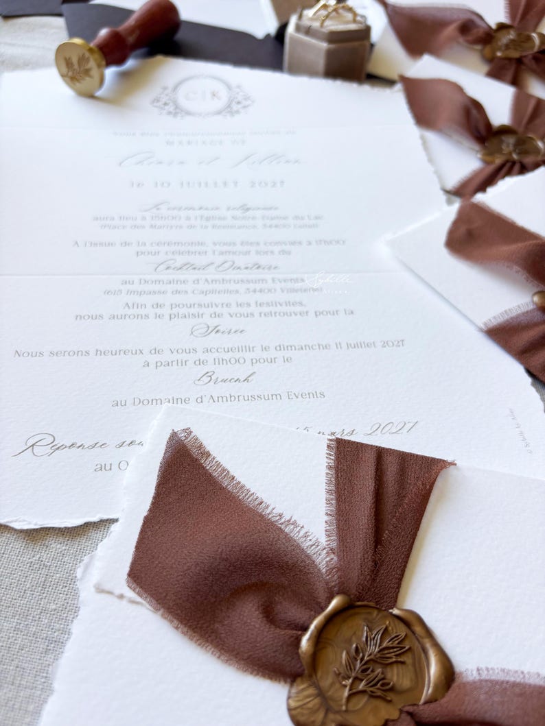 May include: White wedding invitation cards with deckled edges, featuring elegant black script text, brown silk ribbons, and gold wax seals with a botanical leaf design. A wooden wax seal stamp and a small beige ring box are also visible.