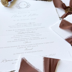 May include: A wedding invitation suite featuring white textured paper with deckled edges, brown silk ribbons, and gold wax seals with a leaf motif. Includes a wooden wax stamp, a beige velvet ring box with two gold rings, and black envelopes.