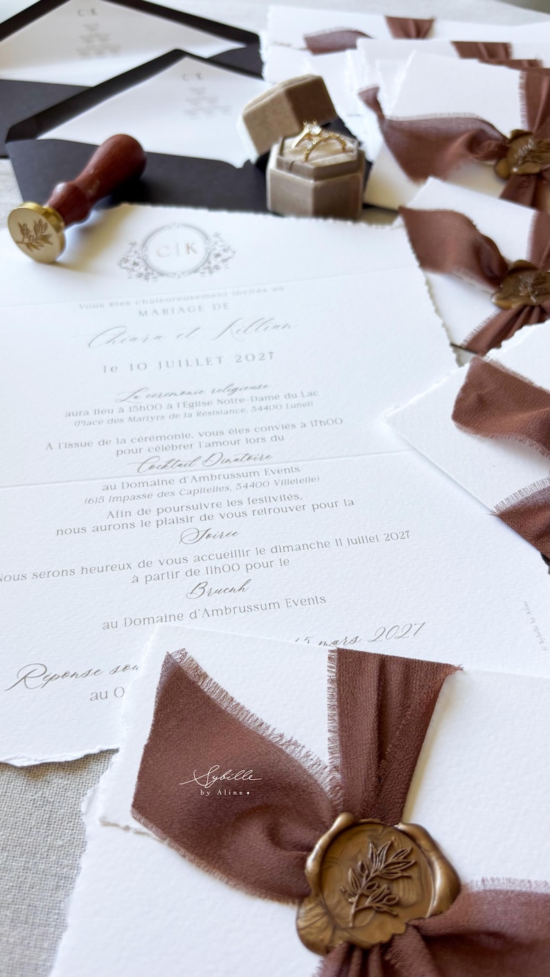 May include: A wedding invitation suite featuring white textured paper with deckled edges, brown silk ribbons, and gold wax seals with a leaf motif. Includes a wooden wax stamp, a beige velvet ring box with two gold rings, and black envelopes.