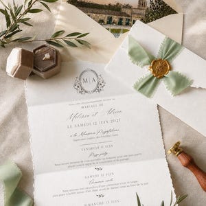 May include: A wedding invitation suite featuring a deckled-edge paper card with black text, a gold wax seal on a sage green ribbon, a matching envelope with a printed landscape, a velvet ring box containing a gold ring, and olive branches.