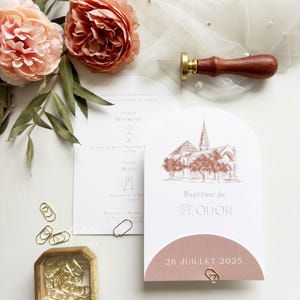 May include: Elegant baptism invitation suite featuring a church illustration in a soft rose hue. Includes a matching envelope, paperclips, and a wooden handled wax seal. The invitation is adorned with floral accents and the date "26 JUILLET 2025".