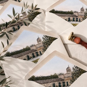 May include: A collection of cream-coloured envelopes with custom envelope liners featuring a watercolour illustration of a stone building with a bell tower, green doors, and string lights, accompanied by a brass wax seal stamp with a wooden handle.