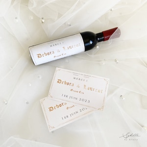 Può includere: Due carte bianche con lettere dorate che dicono "Merci! Debora & Laurent Grand Cru 1er Juin 2023" e "Debora Grand 1er Juin 20". Una bottiglia di vino in miniatura con un tappo rosso e un'etichetta bianca che dice "Merci! Debora & Laurent Grand Cru" è sopra le carte.