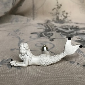 May include: White ceramic mermaid door knob with a black tail fin and a detailed face. The mermaid is lying on her side with her tail curled up.