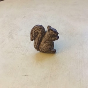 May include: A brown ceramic squirrel figurine with a detailed design. The squirrel is sitting on its haunches with its tail curled up behind it.