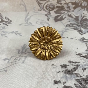 May include: A gold-coloured, flower-shaped decorative knob. The knob features a detailed design with multiple petals and a central floral pattern. The knob is likely made of metal and is set against a patterned fabric background.