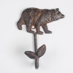 May include: A decorative metal wall hook with a bear motif. The brown bear has a textured finish and sits above a stem with leaf detailing. The hook is suitable for hanging coats or other items.