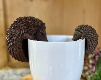 Könnte beinhalten: Zwei braune Keramik-Becherhalter in Form von Igeln, einer auf jeder Seite einer weißen Keramiktasse.