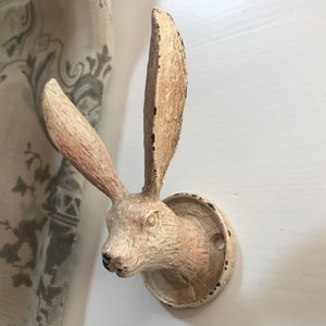 May include: A vintage-style wall hook in the shape of a rabbit's head, made from off-white metal with distressed detailing. The hook features long ears and a circular base. Black accents add to the aged appearance. It is mounted on a white wall.