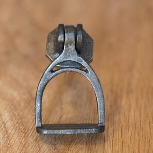 May include: A distressed metal drawer pull in a stirrup shape. The pull has a rectangular top, an arched middle, and a flat base. The metal has an aged, antique finish, giving it a vintage appearance. The measurements are in centimeters.