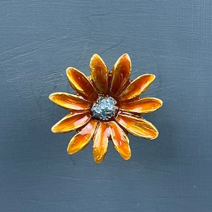 May include: A gold-toned metal flower with orange enamel and a blue rhinestone centre.