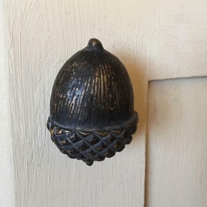 May include: A bronze-coloured acorn-shaped cabinet knob. The knob has a textured surface with vertical lines on the top and a diamond pattern on the bottom. It is mounted on a cream-coloured wooden surface.