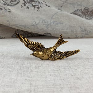 May include: A gold-coloured metal bird figurine in flight. The bird has detailed wing and tail feathers. The figurine is set against a neutral background, highlighting the bird's form and texture. The bird's wings are outstretched.