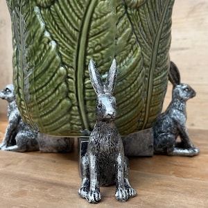 May include: A green ceramic planter with a cabbage leaf design, supported by three silver-coloured rabbit figurines. The rabbits are in a seated position, adding a whimsical touch to the planter's base. The planter is on a wooden surface.