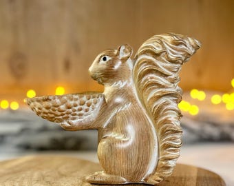 Può includere: Una statuetta di scoiattolo in legno con una coda folta, che tiene una piccola ciotola nelle sue zampe anteriori. Lo scoiattolo è dipinto in tonalità di marrone e bianco, che gli conferiscono un aspetto rustico.