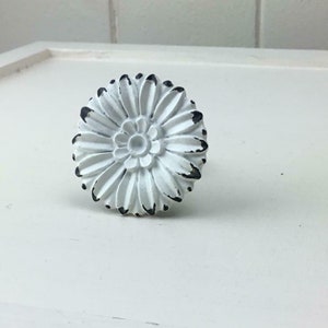 May include: White ceramic flower-shaped knob with a distressed finish. The knob has a raised centre with a smaller flower design.