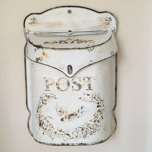 Pu&ograve; includere: Cassetta postale in metallo bianco con finitura usurata e la scritta "POST" in lettere nere. La cassetta postale ha un motivo floreale decorativo e un uccello.