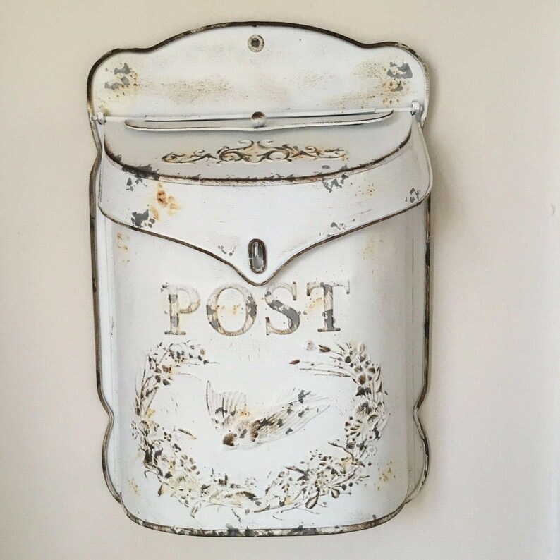 Pu&ograve; includere: Cassetta postale in metallo bianco con finitura usurata e la scritta "POST" in lettere nere. La cassetta postale ha un motivo floreale decorativo e un uccello.