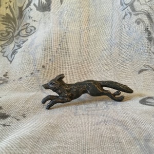 May include: A bronze-coloured metal figurine of a fox running. The fox is in mid-stride with its tail raised and its head turned to the side. The figurine is detailed with fur and facial features.