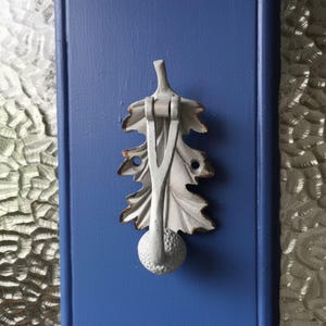 May include: A grey, leaf-shaped door knocker with a textured surface and bronze-coloured edges is mounted on a blue door. The knocker features a detailed oak leaf design and a round, textured base.