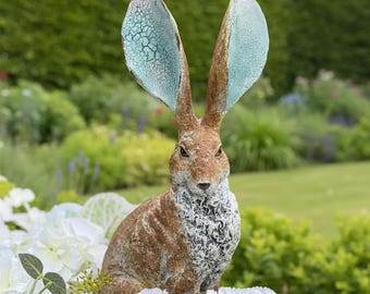 Può includere: Statua da giardino dettagliata di una lepre con lunghe orecchie. La lepre ha un corpo marrone con una superficie strutturata e accenti blu-verdi sulle orecchie. È circondata da fiori bianchi e verde.