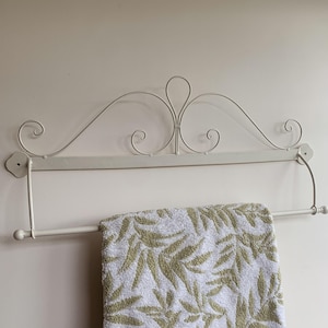 May include: White metal towel rack with a decorative scroll design. The rack has a bar that extends out to hold a towel. A white and green patterned towel is hanging on the rack.