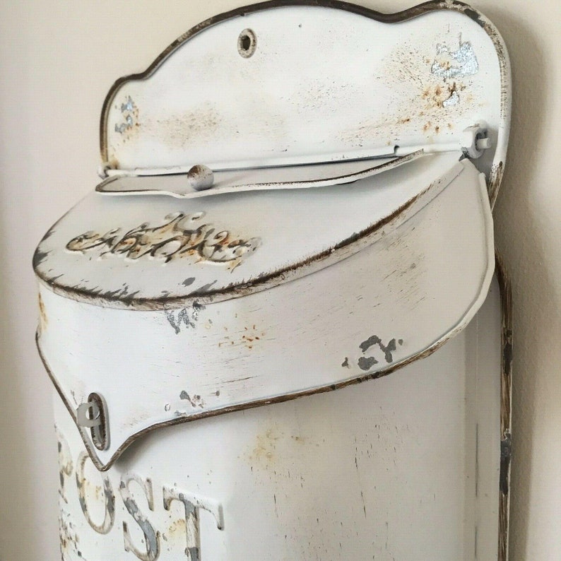 Pu&ograve; includere: Cassetta postale in metallo bianco con finitura usurata e design decorativo. La cassetta postale ha una fessura per la posta e uno sportello che si apre per rivelare l'interno. La cassetta postale &egrave; etichettata "POST".