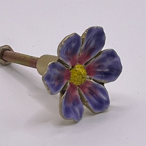 May include: A purple and pink flower-shaped cabinet knob with a yellow centre. The knob is attached to a gold metal screw.