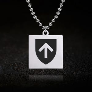 May include: A silver-colored pendant necklace featuring a square pendant with a black shield design and a white arrow pointing upwards. The necklace has a ball chain and is displayed against a dark background.