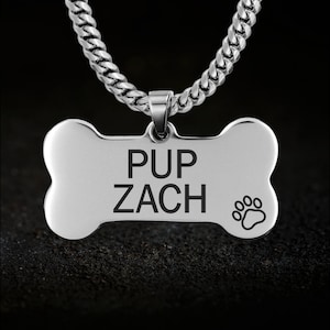 Könnte beinhalten: Ein silberner Hundeanhänger in Knochenform, graviert mit "PUP ZACH" und einem Pfotenabdruck. Der Anhänger hängt an einer silbernen Kette. Der Hintergrund ist dunkel.