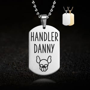 May include: A silver dog tag necklace with the words "HANDLER DANNY" in black, along with a dog graphic. The necklace has a ball chain. Two additional dog tags are visible in the background.