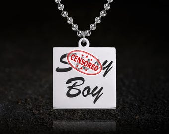 Könnte beinhalten: Silberfarbene Halskette mit quadratischem Anhänger. Der Anhänger trägt die Worte "Sexy Boy" in Schreibschrift, mit einem roten "CENSORED" Stempel über dem Wort "Sexy". Die Halskette hat eine Kette aus kleinen, runden Kugeln.