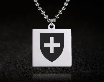 May include: A silver-colored pendant necklace featuring a square pendant with a black shield design and a white cross. The necklace has a ball chain and is displayed against a dark background.