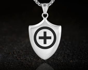 May include: A silver-colored pendant with a shield shape, featuring a black cross within a circle. The pendant hangs from a silver chain. The background is dark, highlighting the pendant's design.