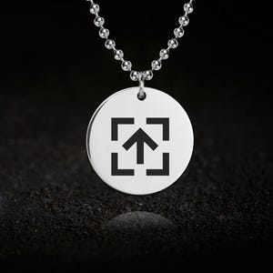 Könnte beinhalten: Eine silberfarbene Halskette mit Anhänger und rundem Charm. Der Charm zeigt einen schwarzen Pfeil, der innerhalb eines quadratischen Rahmens nach oben zeigt. Die Halskette hat eine Kugelkette und ist vor einem dunklen, strukturierten Hintergrund platziert.
