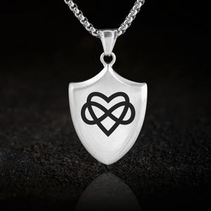May include: Silver-toned pendant necklace featuring a shield-shaped charm. The charm displays a black infinity heart design. The necklace has a chain link style and is displayed against a dark background.