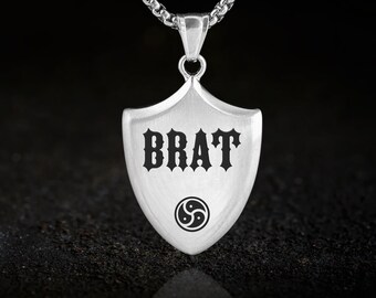 May include: A silver-colored pendant necklace with a shield-shaped charm. The charm features the word "BRAT" in bold, gothic-style lettering and a circular symbol. The necklace chain is a linked design.