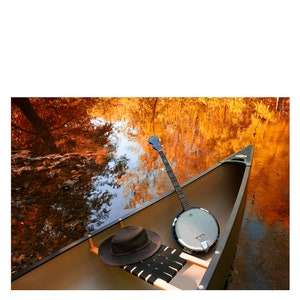 May include: A brown canoe with a banjo and a black hat in it. The canoe is on a calm body of water with a reflection of fall foliage.