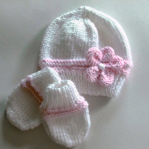 May include: White knitted baby hat with a pink flower and matching mittens. The hat and mittens have pink trim.