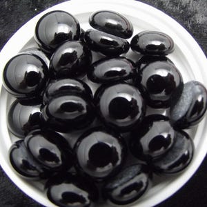 May include: A collection of smooth, black, polished stones of various shapes, including round and oval, are arranged in a white, shallow bowl. The stones have a glossy, reflective surface.
