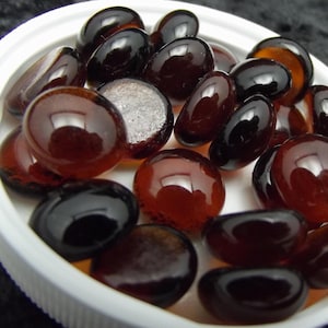 May include: A collection of polished, dark brown glass pebbles in various shapes and sizes, displayed in a white, shallow dish. The pebbles have a glossy, reflective surface, with some appearing nearly black and others a rich amber color.