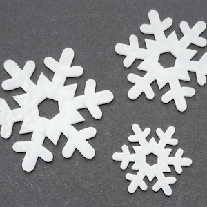 May include: Three white paper snowflakes of varying sizes on a gray background. The snowflakes are cut out and have a delicate, intricate design.