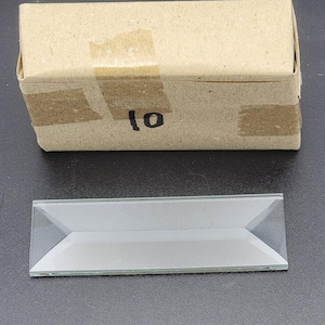 May include: A rectangular glass prism with beveled edges is positioned below a small, brown cardboard box. The box is sealed with tape and has the number "10" written on it in black marker. The prism is clear and reflects light.