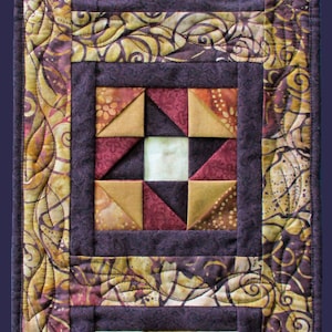 May include: A quilted table runner with a brown, gold, and white geometric pattern. The runner is framed with a dark brown border and a brown, gold, and white floral pattern.