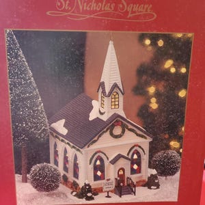May include: A Christmas-themed miniature wedding chapel from The Village Collection by St. Nicholas Square. The white chapel has a dark gray roof, a tall steeple, and colorful stained-glass windows. Snow covers the roof and ground, with a Christmas tree and lights in the background.