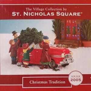 May include: A Christmas-themed figurine depicting a red vintage car with a Christmas tree on top, surrounded by figures. The scene is set in a snowy environment, with a house in the background. The text "Christmas Tradition" is visible.