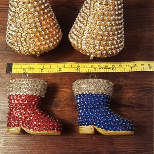 May include: Two gold beaded bell-shaped ornaments with cream-colored ribbon hangers. Below are two boot-shaped ornaments, one red and one blue, both with silver tops and gold soles. A ruler is placed between the ornaments.