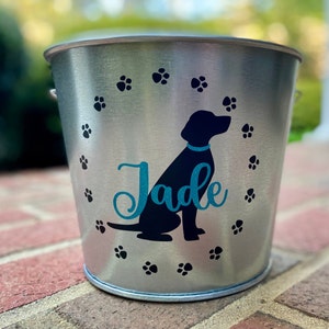 May include: Silver metal dog treat bucket with a black silhouette of a dog and the name "Jade" in teal blue lettering. The bucket is decorated with paw prints.