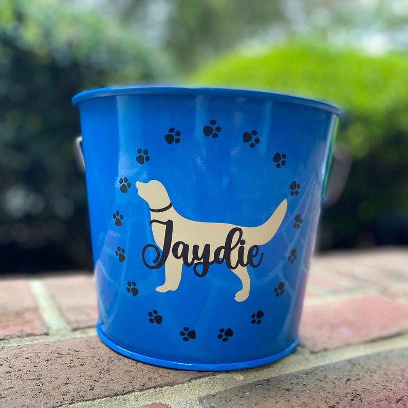 May include: Blue metal bucket with a white dog silhouette and the name "Jaydie" printed on it. The bucket is decorated with black paw prints.