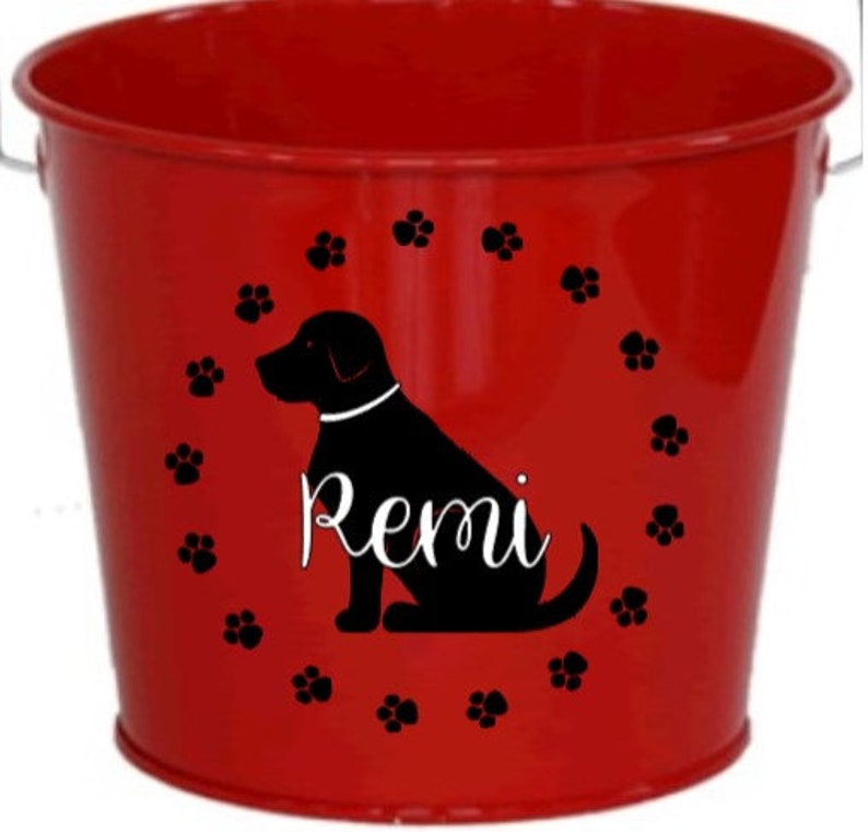 May include: Red metal bucket with a black silhouette of a dog and the name "Remi" in a white cursive font. The dog is surrounded by a circle of black paw prints.